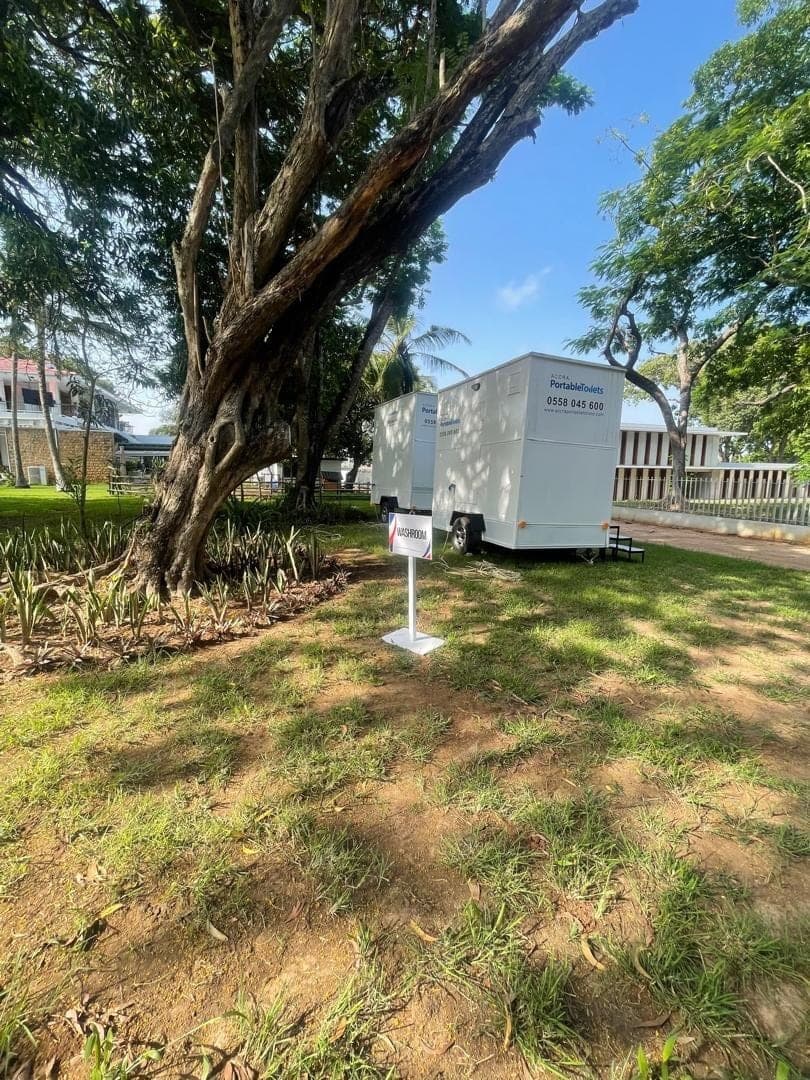 Restroom trailer set up on grass in the shade of a large tree